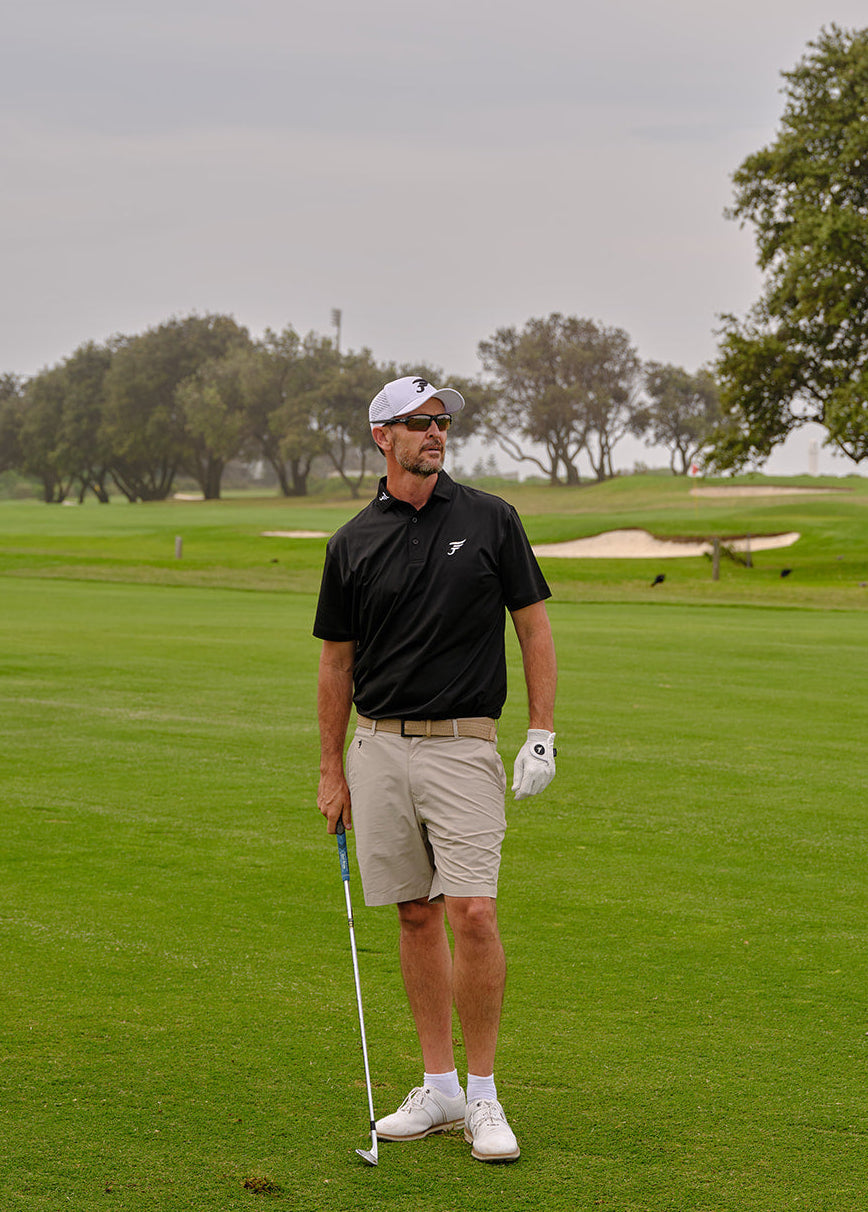 Man on a golf course holding a golf club and ball, with trees and greenery in the background.