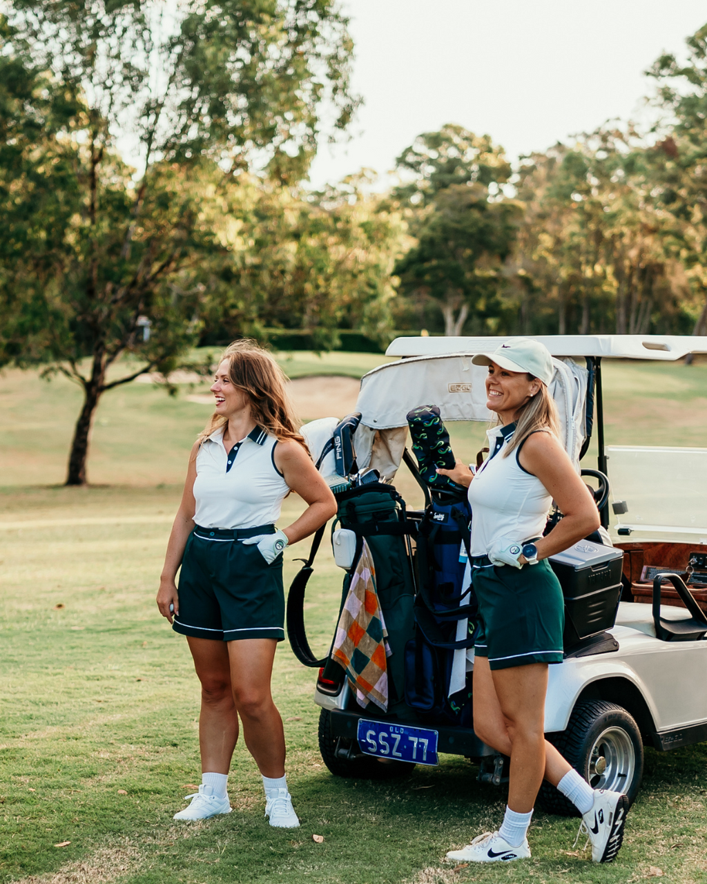 Two women wearing Margie Polo and Stella Tailored Golf Shorts, standing by a golf cart, showcasing stylish, functional golfwear for women.