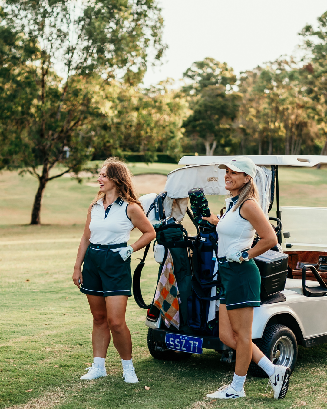 Two women wearing Margie Polo and Stella Tailored Golf Shorts, standing by a golf cart, showcasing stylish, functional golfwear for women.